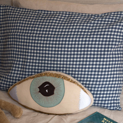 Hazel &amp; George linen pillow in Vichy styled on a bed with a decorative eye-shaped pillow, a plush toy and a book