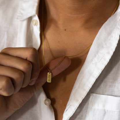 Hazel &amp; George Rectangular Initial Hanger 14K Gold, woman holding a rectangular gold pendant engraved with the letters AMH against a white shirt