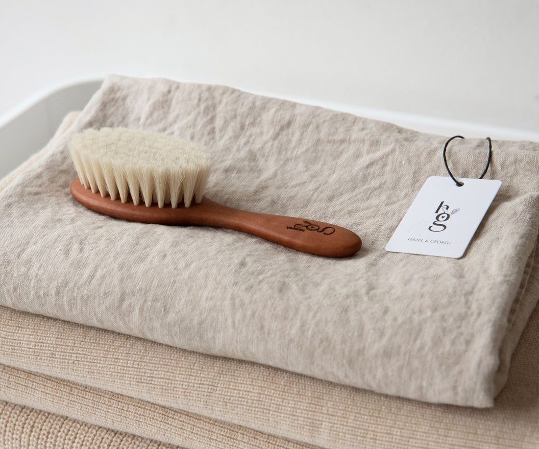 Hazel &amp; George wooden baby hair brush lying on a folded linen blanket next to the Hazel &amp; George hang tag
