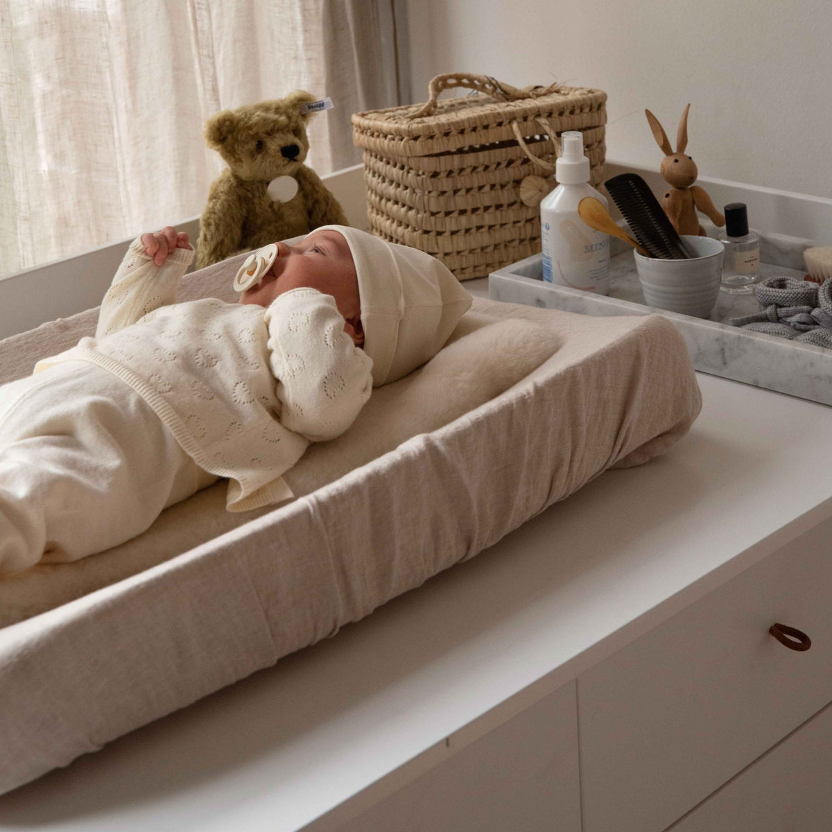 Hazel &amp; George Linen Changing Mat Cover in Craie, with a swaddled newborn lying on the changing pad next to a teddy bear, woven basket and changing table accessories