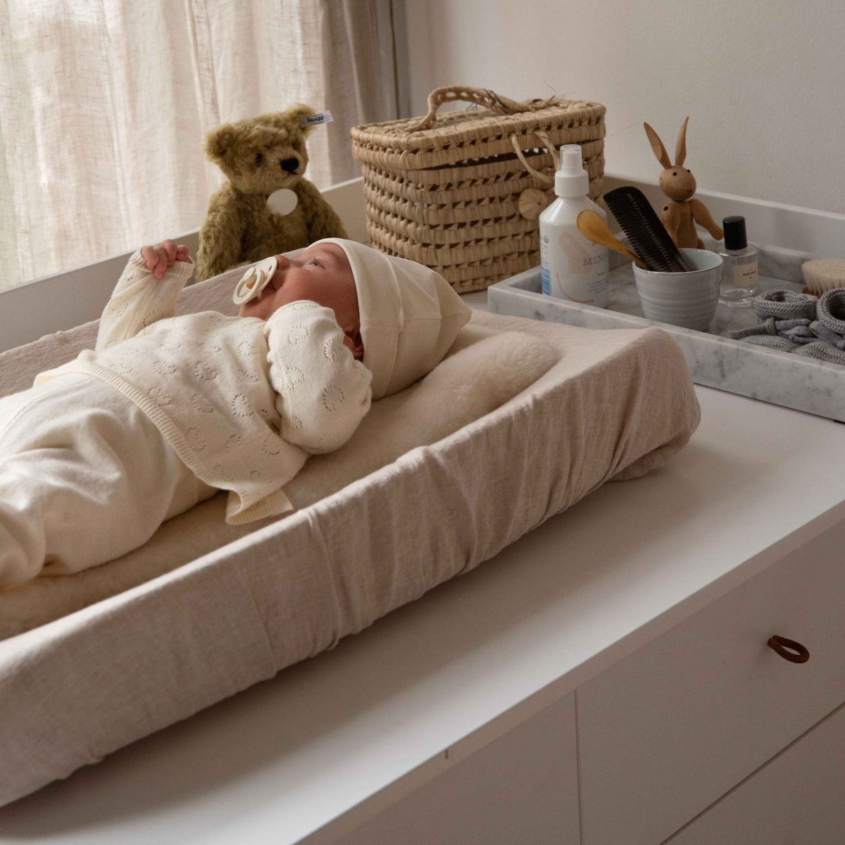 Hazel &amp; George Linen Changing Mat Cover, with a swaddled newborn lying on the changing pad next to a teddy bear, woven basket and changing table accessories