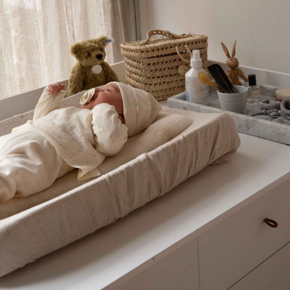 Hazel &amp; George Linen Changing Mat Cover, with a swaddled newborn lying on the changing pad next to a teddy bear, woven basket and changing table accessories