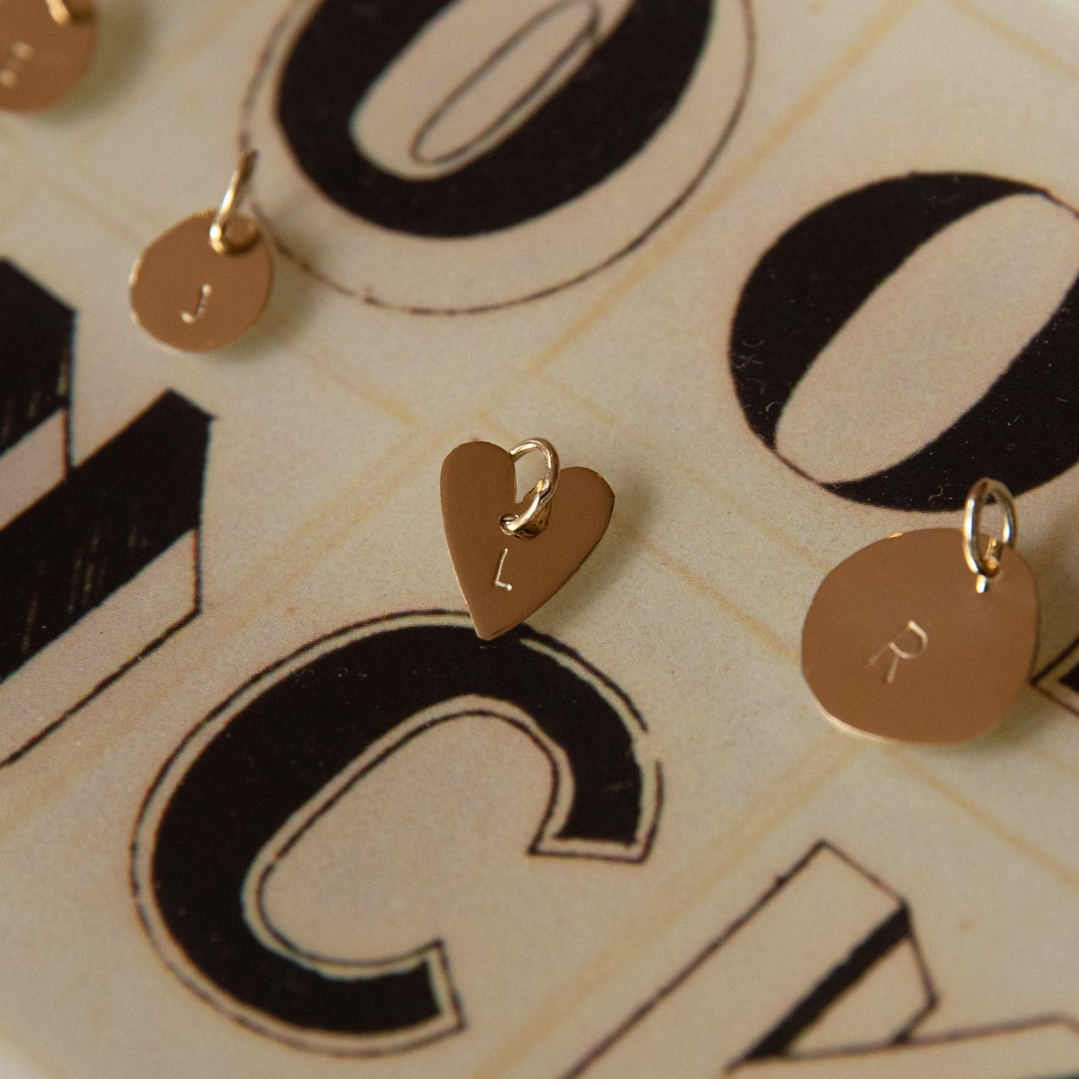 Hazel &amp; George Heart Initial Hanger, gold heart-shaped pendant engraved with the letter L displayed together with small round initial charms, on a graphic letter print background