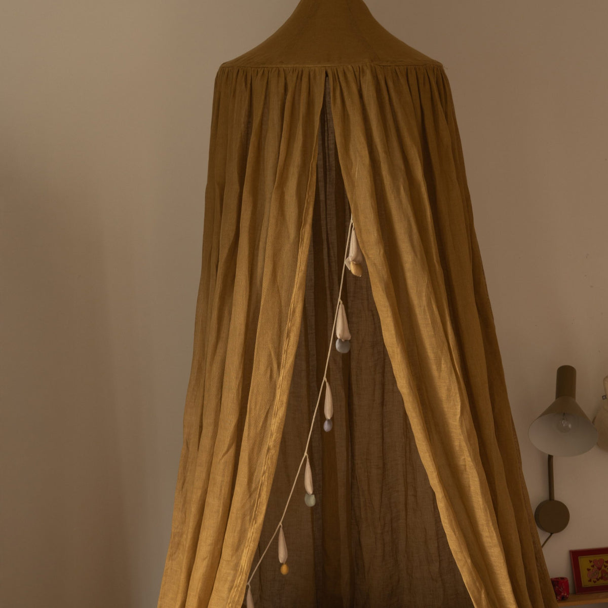 Hazel &amp; George Linen Bed Canopy in Dore in a bedroom corner, with a beaded garland hanging inside and a wall lamp nearby