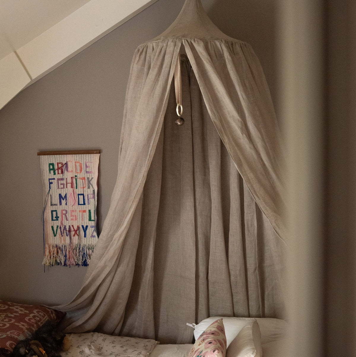 Hazel &amp; George Linen Bed Canopy in Naturel hanging above a bed in an attic bedroom, with an embroidered alphabet wall hanging and floral pillows