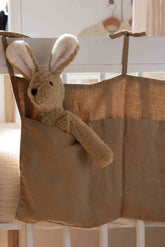 Hazel & George Linen Bed Pocket in Dore tied to a white cot rail, with a plush bunny toy peeking out of the pocket