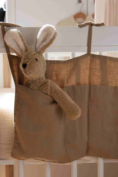 Hazel &amp; George Linen Bed Pocket in Dore tied to a white cot rail, with a plush bunny toy peeking out of the pocket