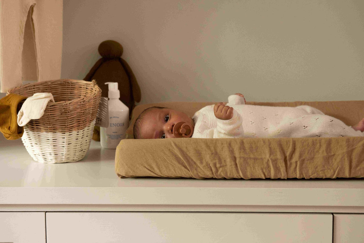 Hazel &amp; George Linen Changing Mat Cover in Dore, with a baby lying on the changing pad on a changing table, next to a basket and plush toy