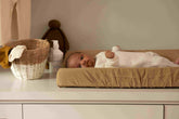Hazel & George Linen Changing Mat Cover in Dore, with a baby lying on the changing pad on a changing table, next to a basket and plush toy