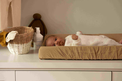 Hazel &amp; George Linen Changing Mat Cover in Dore, with a baby lying on the changing pad on a changing table, next to a basket and plush toy