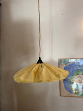A handmade pendant light with a pleated yellow fabric shade and a black metal top, hanging from a rope cord against a beige wall next to a framed picture.