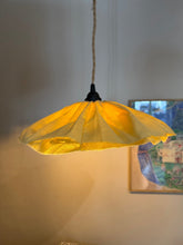 A hanging pendant light with a translucent yellow fabric shade and a natural jute cord, shown lit against a warm beige wall with a framed picture in the background.
