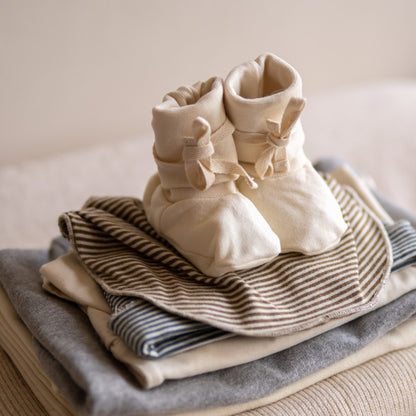 Gray Label Ribbed Baby Booties in Cream, styled on top of folded striped baby clothing